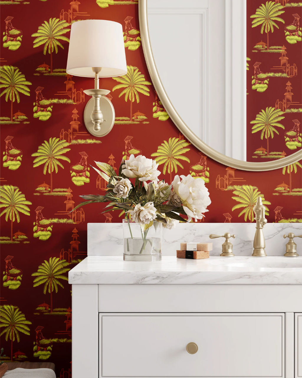 A styled bathroom corner featuring a white vanity with a marble countertop, brass fixtures, a vase of white flowers, a wall-mounted lamp, and a large oval mirror set against a bold red Bali Rice Fields wallpaper with yellow palm tree and architectural motifs.