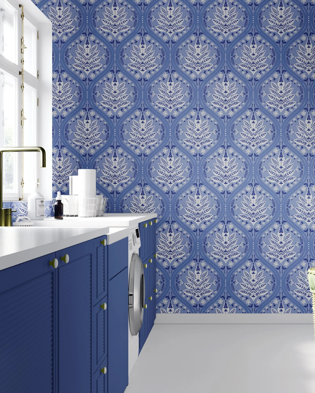 A modern laundry area featuring bold blue and white damask Eden pattern wallpaper, blue cabinets, a white countertop, and a built-in washing machine.