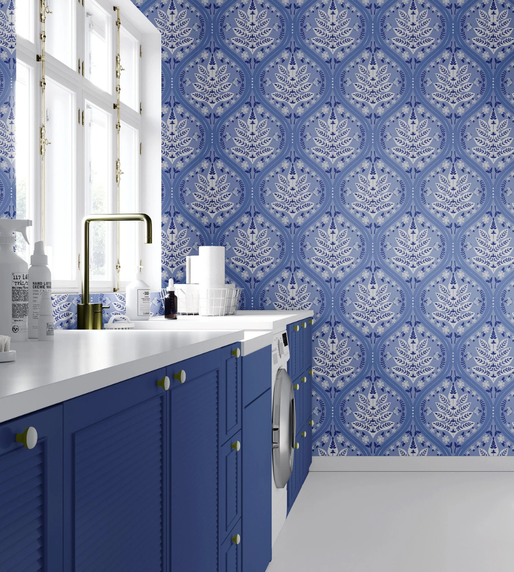 A modern laundry space featuring a bold blue and white Eden pattern wallpaper on the back wall, matching blue cabinets, a white countertop, and a gold faucet.