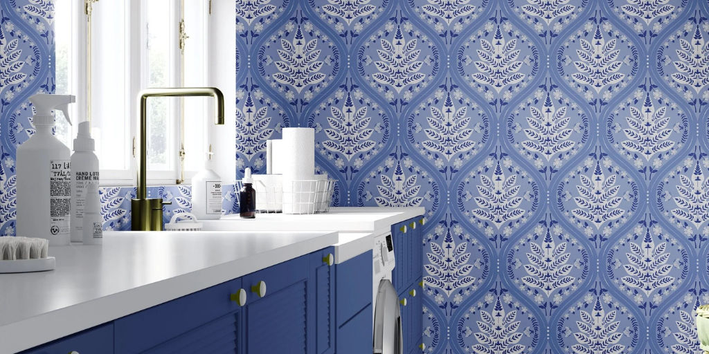 Blue and white Eden patterned wallpaper installed on a laundry room wall, shown in a styled interior with blue cabinets, a white countertop, and a gold faucet.
