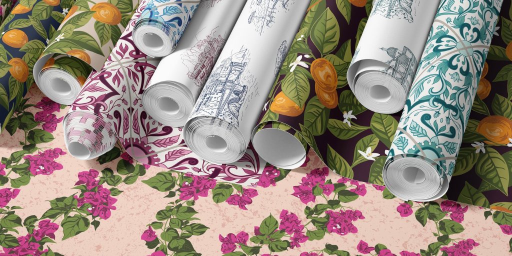 Several rolls of patterned wallpaper for a laundry room arranged diagonally on top of a floral wallpaper sample. The rolls feature white backgrounds with blue architectural illustrations, purple damask motifs, teal floral designs, and a dark botanical print with orange flowers and green leaves.