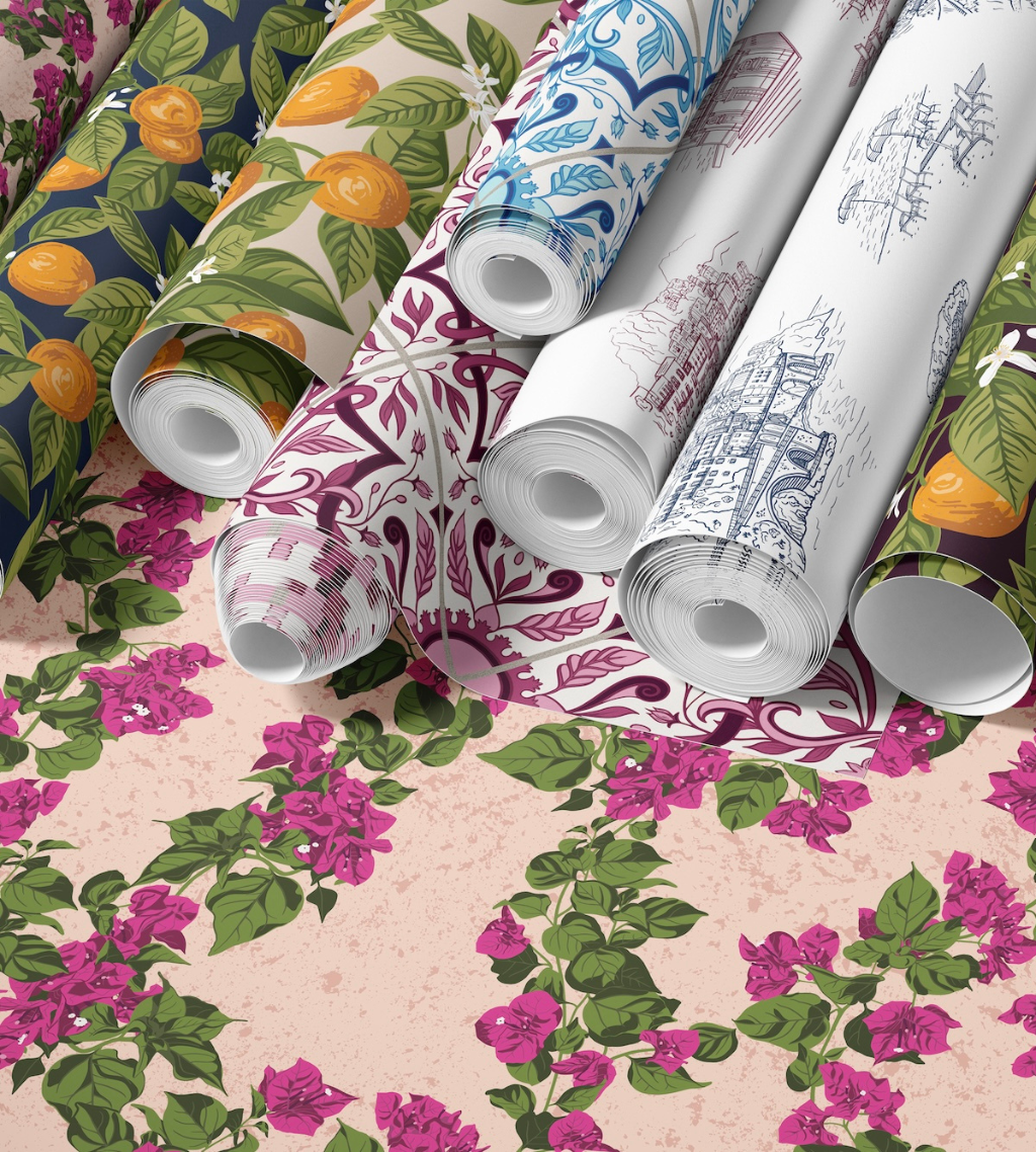 Several rolls of patterned wallpaper for laundry rooms arranged on top of a matching floral wallpaper sample. The rolls show different designs: citrus fruit on a cream background, blue and white ornamental motifs, a white roll with pink floral linework, and a white roll with blue architectural line art.
