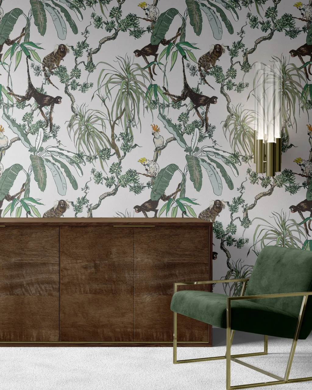 A styled interior scene featuring a wall covered in a detailed botanical and animal White Manila wallpaper with monkeys, tropical leaves, and birds. A dark wood cabinet sits against the wall, and a green velvet armchair with gold metal framing is placed in the foreground.