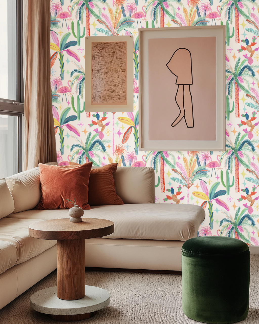 A styled living room corner featuring a beige sectional sofa with rust-orange pillows, a round wooden side table, a dark green velvet ottoman, and a wall covered in colorful Palm Springs Getaway tropical floral wallpaper. Two framed art prints hang on the wall.