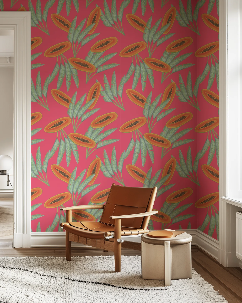 A styled interior room featuring wallpaper with a repeating pattern of green tropical leaves and orange-brown papayas on a bright pink background. A mid-century modern brown leather armchair and a small beige stool sit on a white rug in front of the wall.