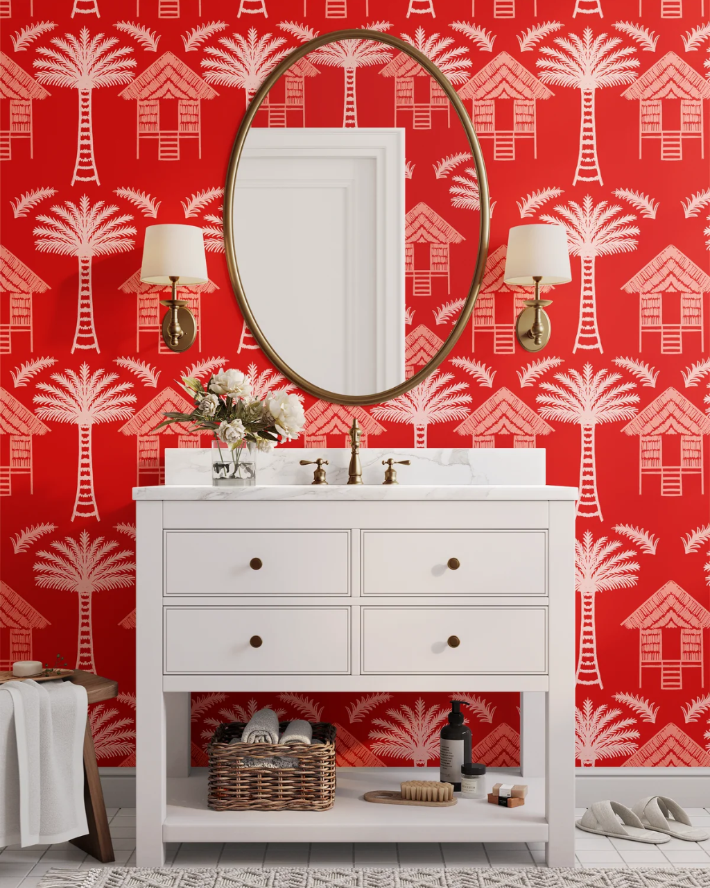 A white bathroom vanity with an open shelf, brass faucet and knobs, set against a bold red tropical huts wallpaper patterned with white palm trees and beach huts. An oval gold-framed mirror hangs above, flanked by two wall sconces.