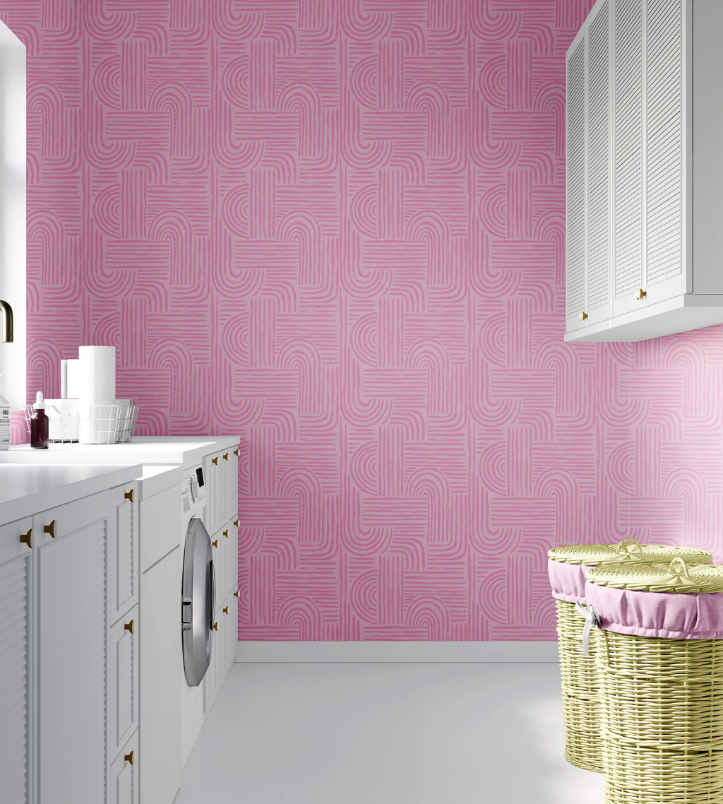 Pink patterned Verona wallpaper on a wall in a bright laundry room with white cabinets, a washing machine, and two wicker baskets with pink fabric liners.