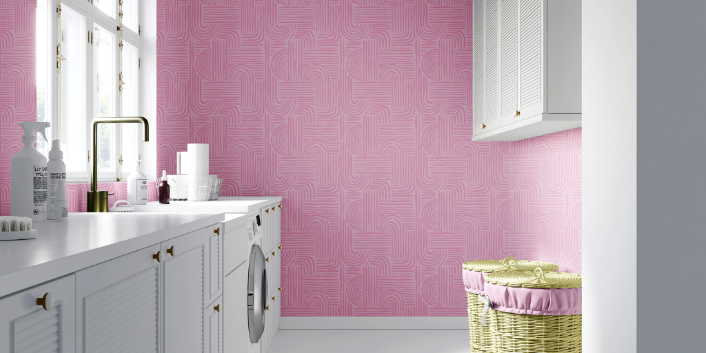 A bright laundry room with pink Verona patterned wallpaper, white cabinets, a sink, and a wicker basket with a pink fabric liner.