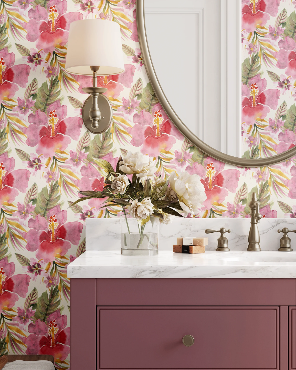 A styled bathroom corner featuring a pink and red floral Watercolor Hibiscus wallpaper with green leaves, a white marble sink countertop, a dusty mauve purple vanity cabinet, a round wall mirror, and a wall-mounted sconce.